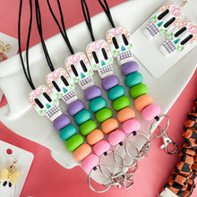 Load image into Gallery viewer, A styled flat-lay of colorful Day of the Dead themed jewelry, featuring sugar skull necklaces with pastel beads and matching skull earrings on white display cards against a pink background.